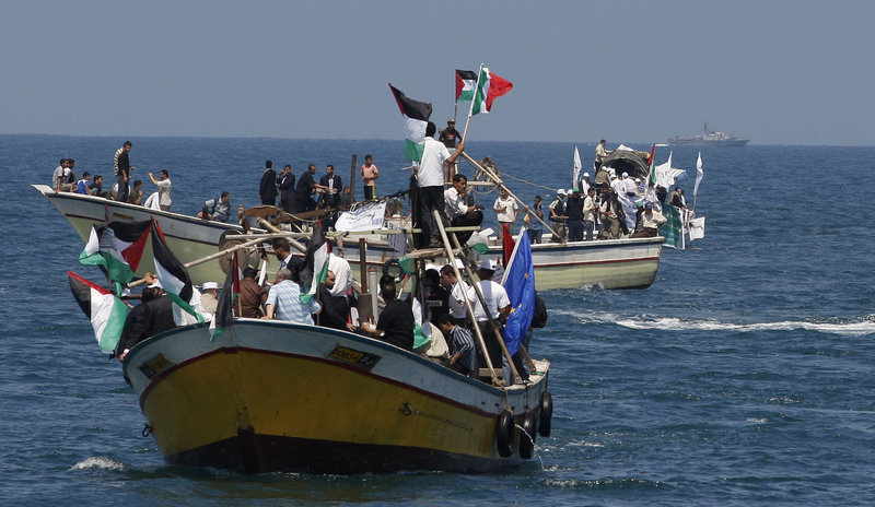 Palestinians ride boats in Gaza waters while an Israeli navy vessel, seen in the background, patrols at a distance. The flotilla of vessels is intent on delivering humanitarian aid to Gaza, while Israel insists the vessels turn back or face “necessary measures’ to enforce the blockade.
