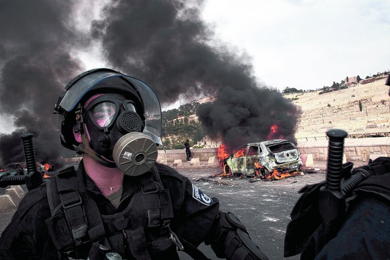 An Israeli police officer wearing a gas mask walks past a car set on fire by Palestinian rioters Wednesday outside Jeru-salem’s Old City. Violence erupted after a Palestinian laborer was killed by a security guard watching over Jewish families.