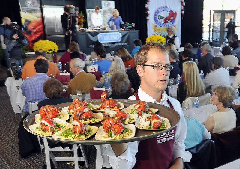 Justin Dewalt carries a tray laden with servings of the winning lobster dish created by Kelly Patrick Farrin, seen on the stage, in the Lobster Chef of the Year competition at Portland’s Ocean Gateway on Friday. Farrin is chef at the Azure Cafe in Freeport.