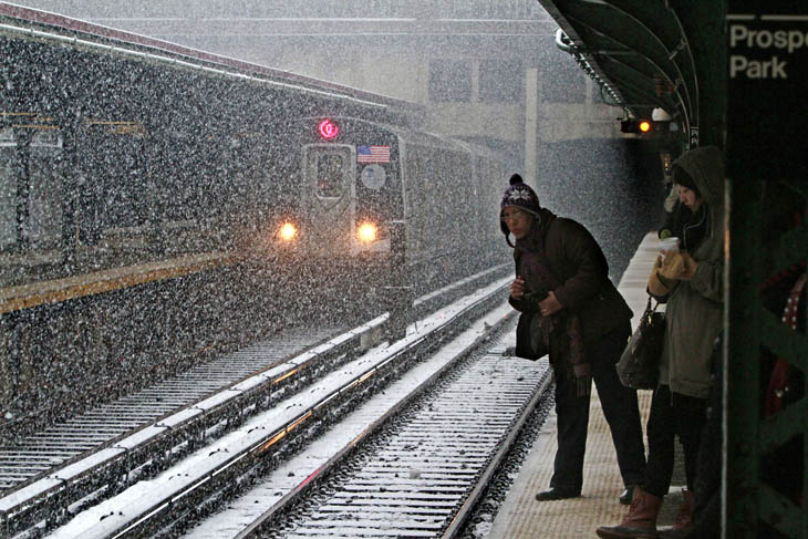A subway train exits a tunnel into snowfall as commuters wait at a station today in Brooklyn, N.Y.