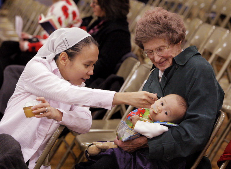 Myra Turmenne, left, feeds applesauce to her 10-month-old daughter Ruth, who is being held by her grandmother, Murielle Turmenne, before the start of a naturalization ceremony at Falmouth Middle School on Thursday. Myra, who is from the Philippines and married to Murielle’s son David, was sworn in as a new citizen at the ceremony.