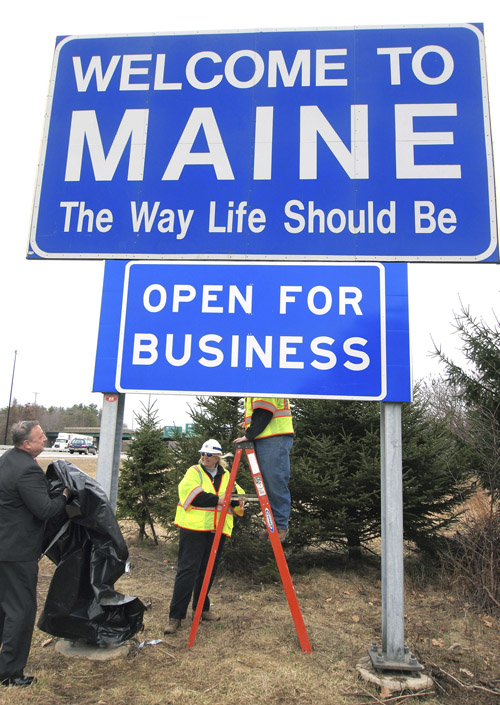 Gov. Paul LePage, left, unveils a new "Open for Business" sign beneath the "Welcome to Maine" sign along Interstate 95 near the New Hampshire border in Kittery today. Maine Department of Transportation crew supervisors Elaine Cota, center, and Aaron Main, on ladder, assisted.