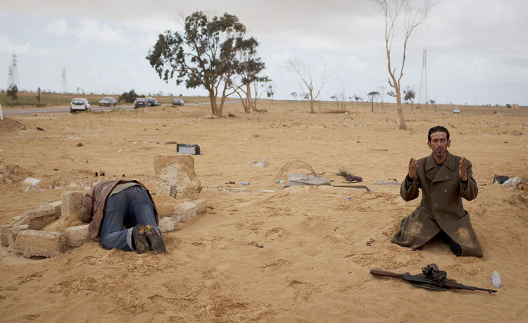 A Libyan rebel prays next to his gun while another kneels over the grave of his dead brother, killed in the fighting, on the outskirts of the city of Ajdabiya, south of Benghazi, Libya, today.