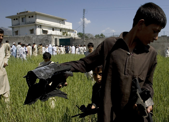 A Pakistani youngster shows metal pieces collected today from a wheat field outside the house, in background, where al-Qaida leader Osama bin Laden was killed in Abbottabad, Pakistan. Local residents showed off small parts of what appeared to be a U.S. helicopter that Washington said malfunctioned and was disabled by the American commando strike team as they retreated.