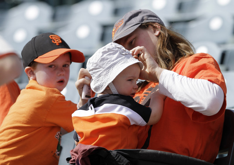 Erica Fortescue, right, of San Francisco applies sunscreen on her 11-month-old daughter Nico during an Oakland Athletics baseball game. At left is Nico’s brother Theo, 4.