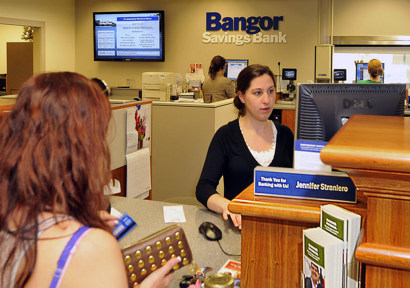 Jennifer Straniero, a teller at the Hammond Street branch of Bangor Savings Bank, serves a customer. On the wall behind her, a flat-screen monitor announces news of supported events in the community.