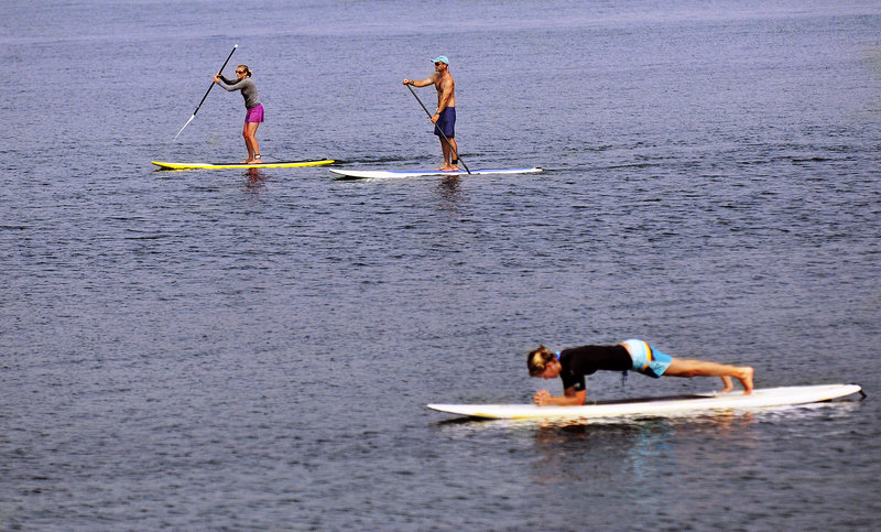 Rafael Adams and Tonya DiMillo pass a fellow paddle boarder practicing yoga on her board in Kettle Cove. “You can paddle for hours with no waves,” Adams said.