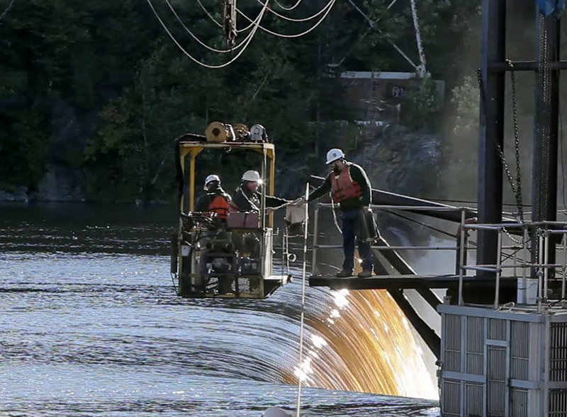 Tyler Grant hands boots to Mitch Simpson before returning to shore in a cable car with Kevin Gallant at Veazie Dam on the Penobscot River in Veazie. The Department of Marine Resources workers were checking for salmon in a cage, at the bottom right, which is at the top of a fishway.