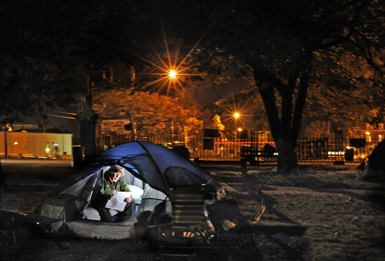 Occupy Maine participant Robert W. Davis IV, also known as “Bobby D,” of Portland, reads a newspaper inside his tent in Lincoln Park on Tuesday night. Davis’ tent, which includes a small mattress, nightstand, lamp and other personal effects, is one of about three dozen that have created a small tent city housing Maine’s incarnation of the protest movement.