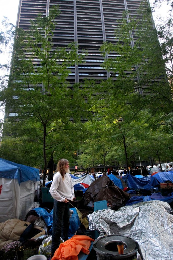 An Occupy Wall Street protester in Zuccotti Park stands amid tarps and sleeping bags Thursday in the shadow of a building owned by Brookfield Properties. Zuccotti Park is owned by a wealthy real estate magnate who has properties worldwide and millions of dollars on hand – precisely the sort of corporation the protesters have been shouting about.
