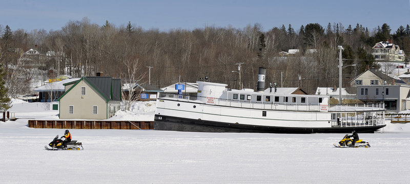 Snowmobilers pass the Katahdin, moored for the winter next to the Moosehead Marine Museum, at left, in Greenville. Bubblers keep the lake water from freezing around the boat’s hull.