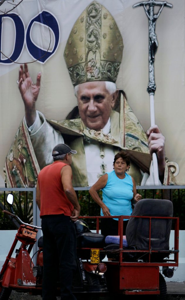 A couple speaks in the street where an image of Pope Benedict XVI hangs in Havana, Cuba, on Thursday. The pope is visiting Mexico and heads for Cuba on Monday.