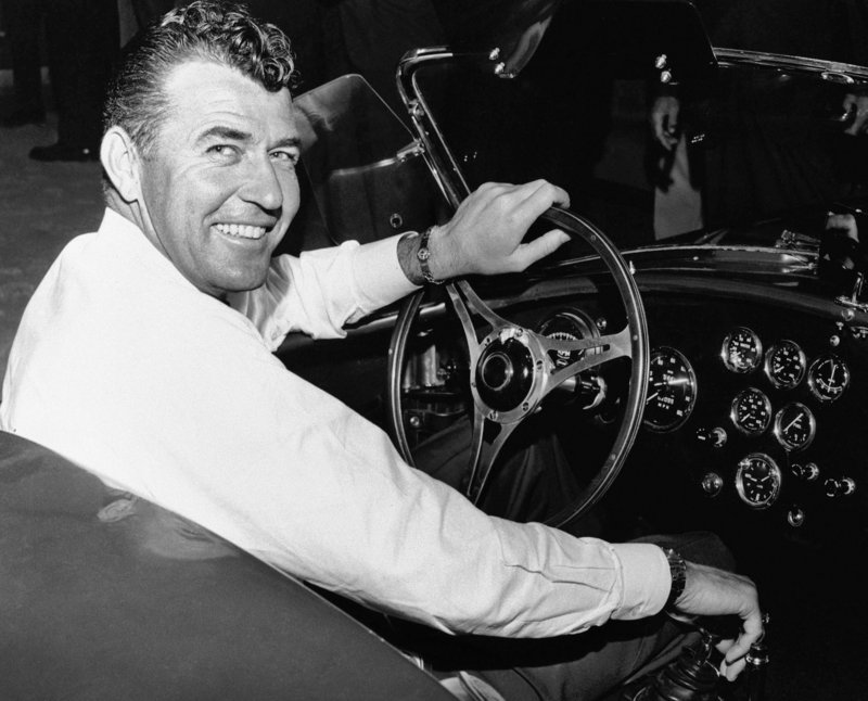 In a 1964 photo, auto racer Carroll Shelby strikes the familiar pose of a car enthusiast behind the wheel. Shelby got into auto racing after his chicken farming business failed when he lost his second flock of chickens to disease.
