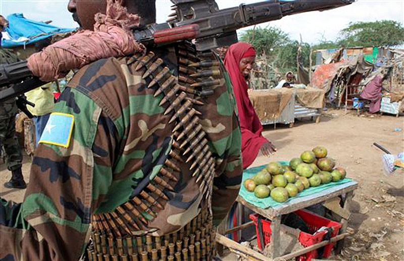In this photo taken Thursday, June 7, 2012, a fruit seller looks across as a Somali government soldier stands guard in Afgoye, west of the capital Mogadishu, in Somalia. The seaside Somali capital is enjoying a peace that, except for the infrequent attack, has lasted the better part of a year and Somalis who fled decades of war are coming back, as are U.N. workers who long operated out of Nairobi, the capital of neighboring Kenya. (AP Photo/Jason Straziuso)