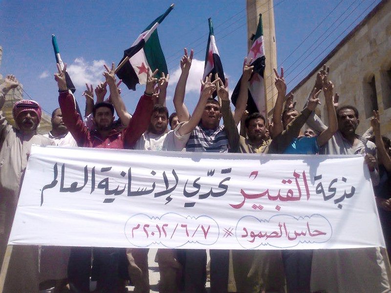In an image provided by Edlib News Network, opponents of the Syrian regime chant slogans Thursday and hold a banner in Arabic that reads, “Al-Qubair massacre challenges the world’s humanity” in the northern village of Hass. There were reports of up to 80 people killed in al-Qubair, but Syrian officials called the reports “absolutely baseless.”