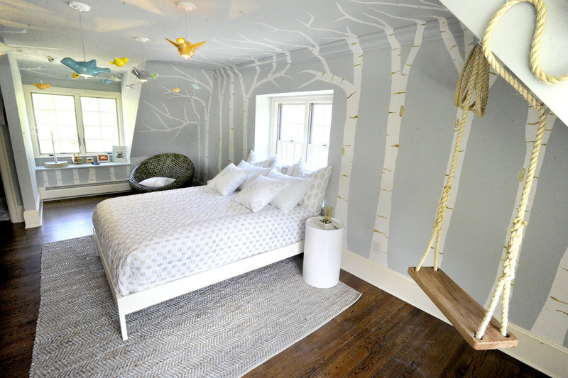 A child’s bedroom, decorated by Nicki Bongiorno of Spaces Kennebunkport, features white birch trees painted on the walls and ceiling, hanging light fixtures shaped like birds, and a rope swing.
