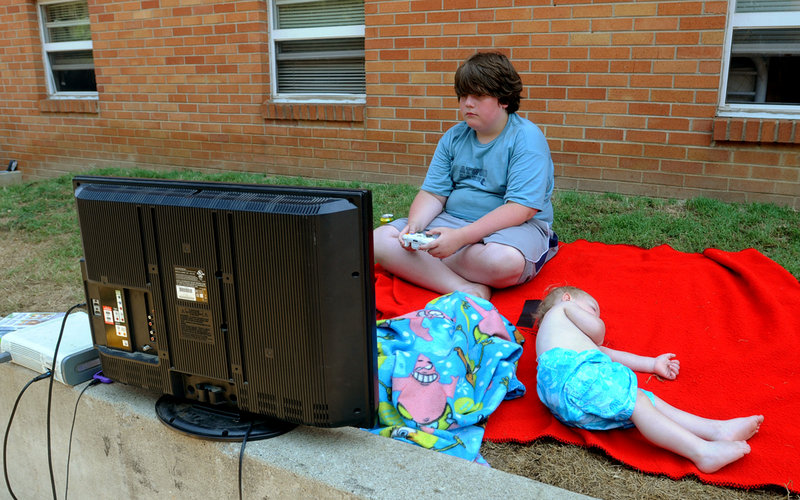 Derek Longo, 11, plays a video game, powered by a running car engine hooked up to a power inverter, outside his home at Bruce Apartments in Ashland, Ky. Sleeping on the ground next to him Monday as they wait for the power to be restored is Joseph DeLong.