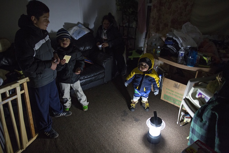 Living through another night of possibly freezing temperatures, Michael Pineda, fifteen months old, stands bundled up near a battery operated lantern in his home without power or heat in the Rockaway Park neighborhood in the Queens borough of New York, Thursday, Nov. 8, 2012, in the wake of Superstorm Sandy. From left is his brother Mario Pineda, 12, Walter Rivera, 5, and center in deep shadow is their mother Fatima Pineda. (AP Photo/Craig Ruttle)