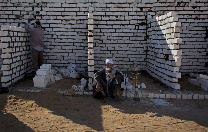 An Egyptian laborer works as his employer smokes a water pipe at the local cemetery that is spreading toward Egypt’s first pyramids and temples.