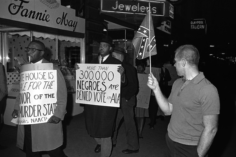 In this April 14, 1964 black-and-white file photo, a man holds a Confederate flag at right, as demonstrators, including one carrying a sign saying: "More than 300,000 Negroes are Denied Vote in Ala", demonstrate in front of an Indianapolis hotel where then-Alabama Governor George Wallace was staying. After more than a century, the Census Bureau is dropping use of the word "Negro" to describe black Americans in its surveys. Instead of the term popularized during the Jim Crow era of racial segregation, census forms will use the more modern-day labels, “black” or “African-American”. (AP Photo/Bob Daugherty, File) Walking Holding Placard Text Flag Social Issues Protestor