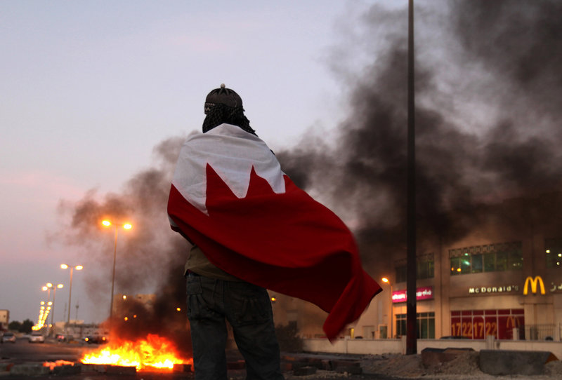 A Bahraini anti-government protester wrapped in a national flag watches tires burn Tuesday in Malkiya, Bahrain. Activists are calling for protesters to flood the streets Wednesday.
