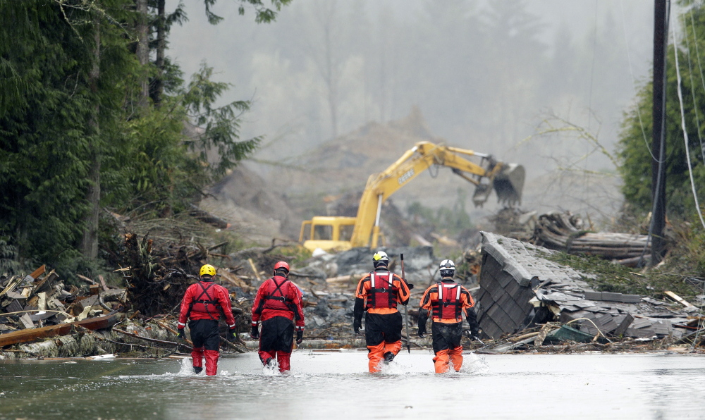 Four search and rescue workers wade through water covering Washington Highway 530 Thursday on the eastern edge of the massive mudslide that struck Saturday near Darrington, Wash. Heavy equipment moves trees and other debris in the background.