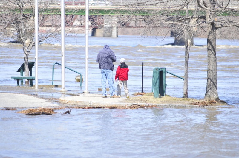 People stand beside the high waters of the Kennebec River near the sign for the Augusta Waterfront Park on Wednesday in downtown Augusta.