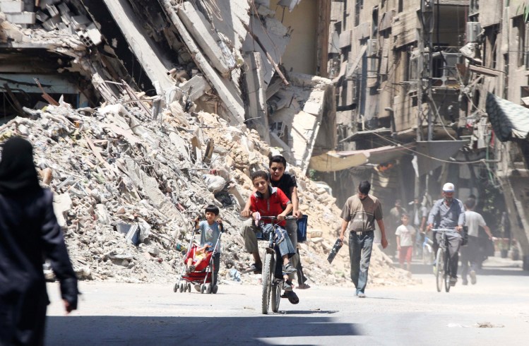 Boys ride a bicycle past other civilians near damaged buildings in the Damascus suburb of Harasta, Syria, on Sunday.