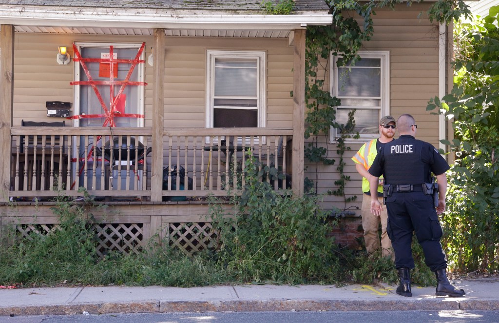 A Blackstone police officer talks with a utility worker while standing post outside a house where a Massachusetts prosecutor said the bodies of three infants were found Thursday. Both "condemned" and "keep out" signs are attached behind police tape to the front door. The utility worker laid some paint markings on the street in front of the house.