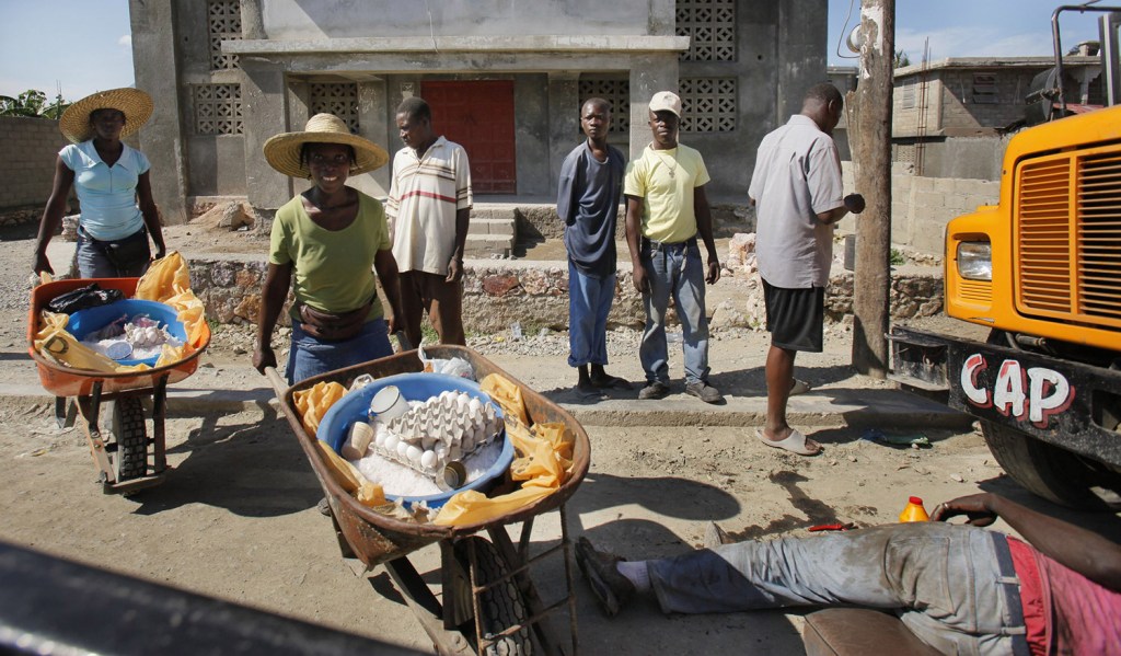 A man works under a bus while two women pushing wheelbarrows of goods wait to pass in Cap Haitien.
