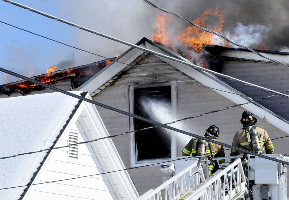 Firefighters spray water into the third-floor window as flames break through the roof of an apartment building on Paris Street in Waterville on Monday. 