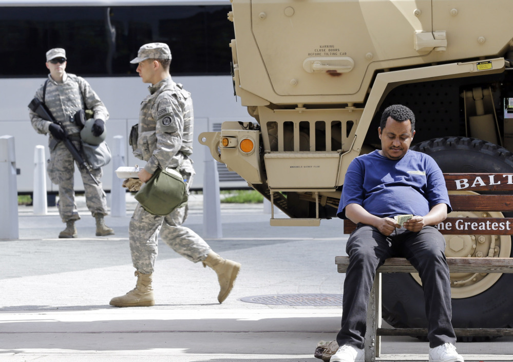 A man sits on a bus stop bench as members of the Maryland National Guard patrol behind him Saturday in Baltimore. The Associated Press