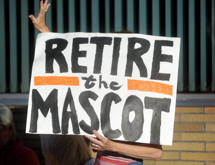 A woman holding a sign urging Skowhegan to change the mascot at the local high school waves on Thursday to a supporter in Skowhegan.