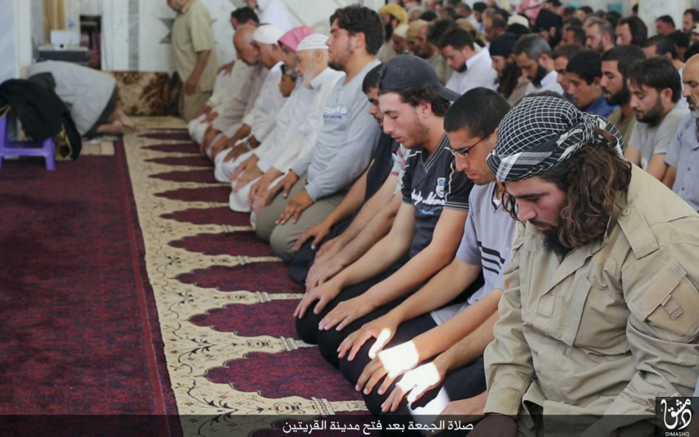 In this photo provided Friday, Aug. 7, 2015, by the Rased News Network, a Facebook page affiliated with Islamic State militants, Muslim worshipers attend Friday prayers in a mosque in the central Syrian town of Qaryatain. The Arabic on the bottom banner reads, “Friday prayers after the conquest of Qaryatain.” Activists on Saturday said hundreds of families fled the Christian town of Sadad as Islamic State militants captured Qaryatain on Thursday, which is about 25 kilometers (15 miles) northwest of Sadad. (Rased News Network, a Facebook page affiliated with Islamic State militants via AP)