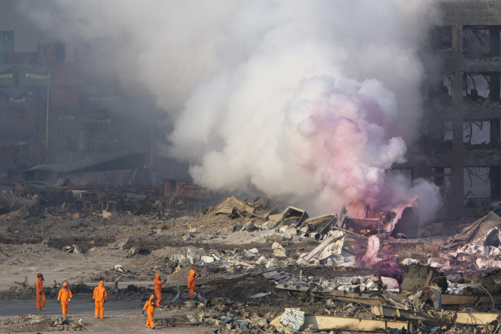 Firefighters in protective gear watch partially pink smoke billow after Wednesday’s explosion. Huge, fiery blasts at a warehouse for hazardous chemicals killed at least 50 people and turned nearby buildings into skeletal shells, raising questions Thursday about whether the materials had been properly stored.