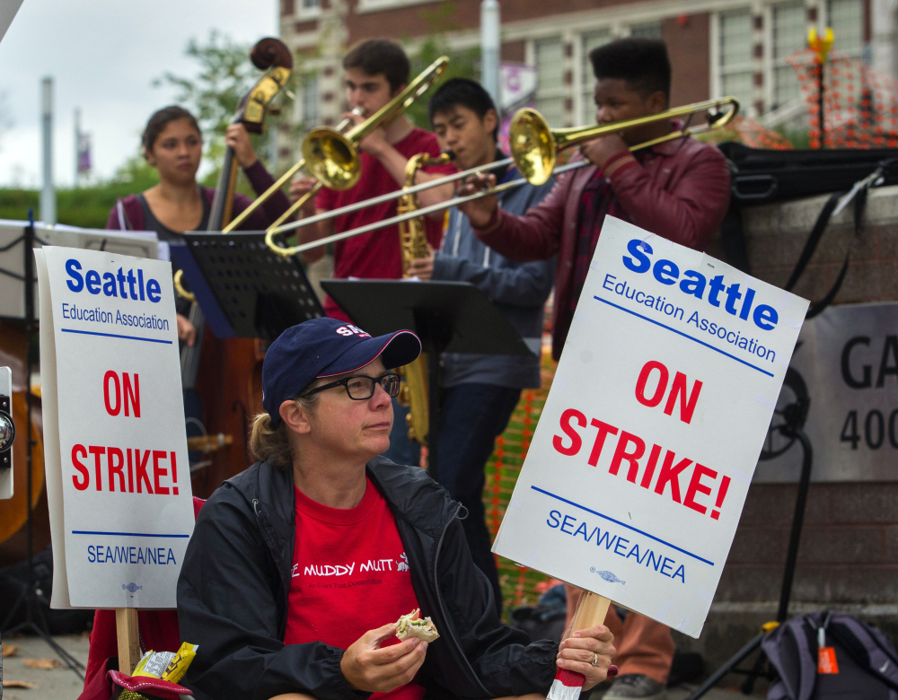Kris McBride, a high school worker, waits to push a traffic-signal button so picketing Seattle teachers can cross the street Monday as student band members play in support.