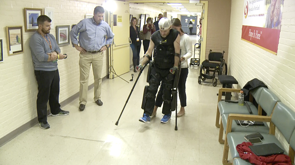 Lauren Peltier of ReWalk Robotics guides Brian Walker at New England Rehabilitation Hospital as he tries out a ReWalk motorized exoskeleton for the first time in Maine, watched by hospital staffers. A small backpack holds the batteries that power the exoskeleton.