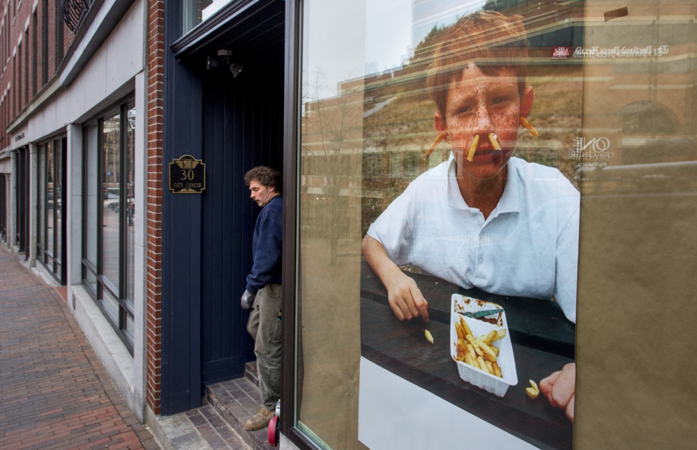 Sam Hopkins of MJ Plumbing & Heating walks past a large poster of a boy with french fries shoved in his ears and nose pasted in the window of what will be Hero, a sandwich shop at 30 City Center. 