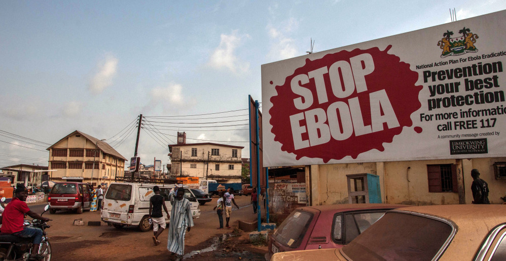 People pass a banner reading “STOP EBOLA,” forming part of Sierra Leone’s Ebola-free campaign in the city of  Freetown, Sierra Leone, on Friday.