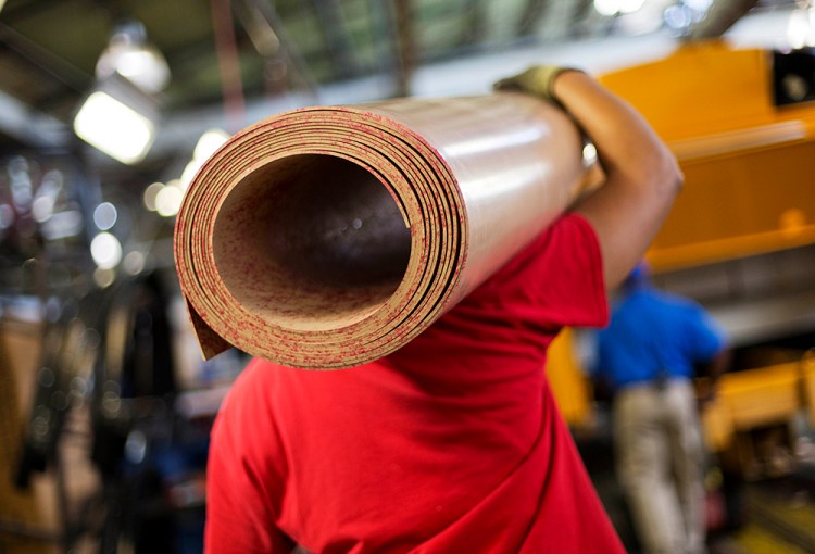 A worker carries a roll of sub flooring to be installed in a school bus on an assembly line at Blue Bird Corp.'s manufacturing facility, in Fort Valley, Ga. Some businesses said that efforts to reduce stockpiles of goods have also held back orders. The Associated Press