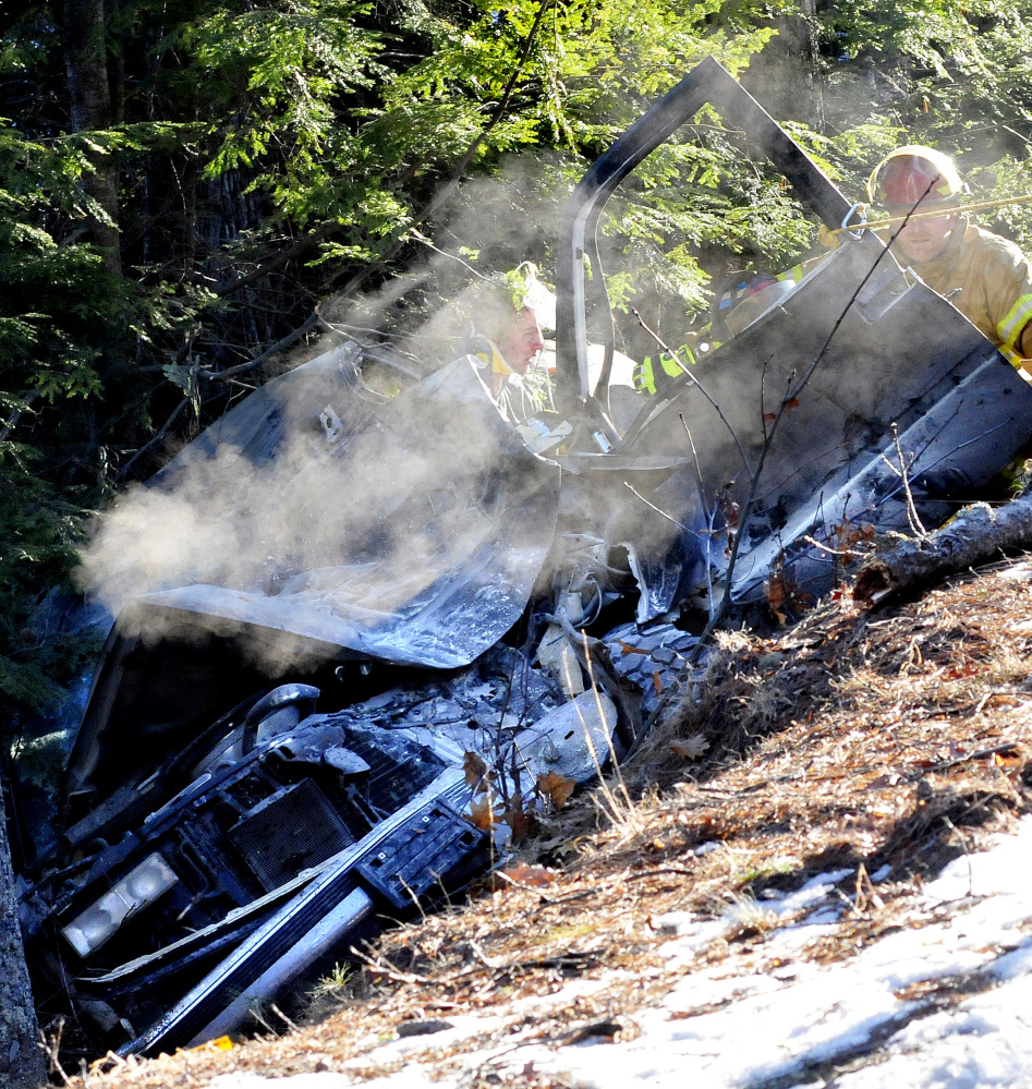 Smoke continues to rise from Eric Bachand’s vehicle as firefighters stabilize him after his vehicle went off Route 135 in Belgrade on Tuesday. A Maine State Police trooper used a fire extinguisher to put out the fire before firefighters arrived.