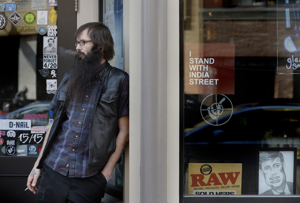 Joshua LeClair, shop manager at The Higher Concept on Congress Street in Portland, takes a cigarette break outside the store Monday. The store is one of many businesses in Portland displaying an "I stand with India Street" sign as they oppose a plan to close the India Street Public Health Center.