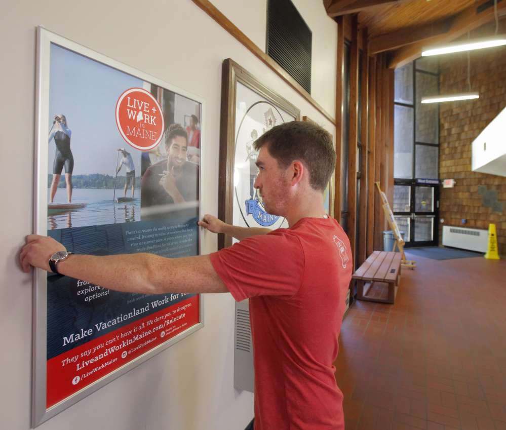 Nate Wildes of Live + Work in Maine puts up a poster at the visitors' center in Kittery. Posters also have been installed at the Portland International Jetport to tout the program.