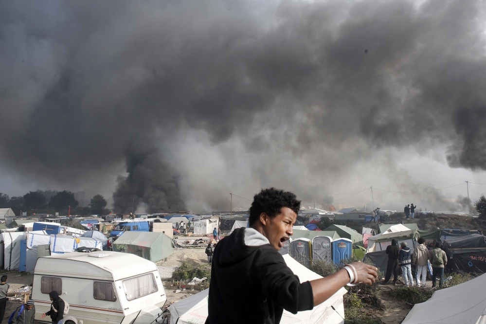A migrant reacts as smoke billows from burning shelters set on fire in the makeshift migrant camp known as "the jungle" near Calais, northern France, Wednesday, Oct. 26, 2016. Firefighters have doused several dozen fires set by migrants as they left the makeshift camp where they have been living near the northern French city of Calais. (AP Photo/Thibault Camus)