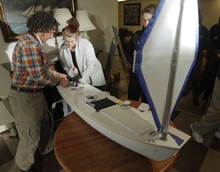 Teacher Ed Sharood, left, and junior Kristin Cofferen, of Kennebunk High School's Alternative Education Class, apply stickers from local businesses on a small boat that they hope will reach Ireland after it is launched. At right, behind the sail, is senior Josh Ellis, who was the boat project manager.