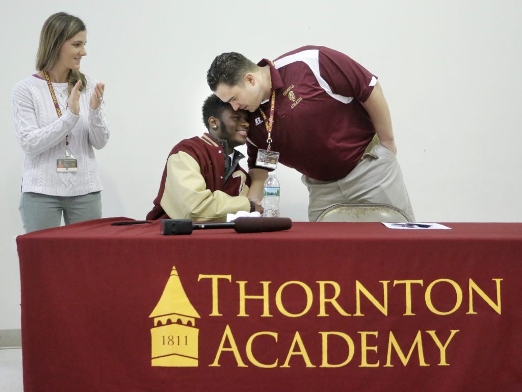 Johnny Rosario is congratulated by Nick Tabor, the offensive line coach for the Thornton Academy football team, after Rosario signed a letter of intent on Wednesday morning at Thornton Academy to play football for the University of Maine. Tabor, along with his wife Danielle, at left, is also a dorm parent to students, including Rosario, in Thornton's residential student program.