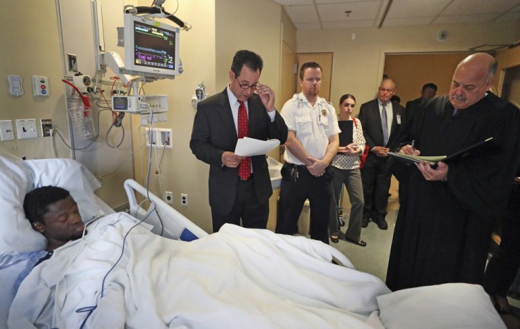 At Tufts Medical Center in Boston, prosecutor John Pappas, second from left, reads as Judge Michael Bolden, right, takes notes during the arraignment of Bampumim Teixeira, who lies in a hospital bed Monday. Teixeira is accused of murdering two engaged doctors in their South Boston penthouse condominium.