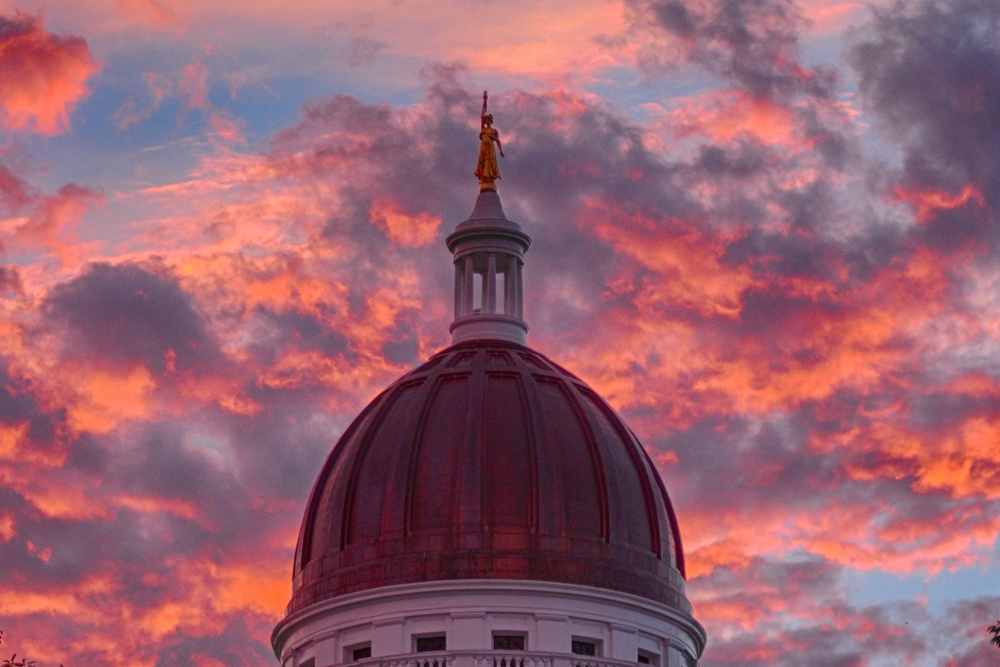 Clouds glow behind the Lady of Wisdom statue on top of the Maine State House dome during a sunset last summer. Cross-party discussions are happening inside as legislators prepare for the inevitable fight over the two-year budget that is required to be passed by July 1.