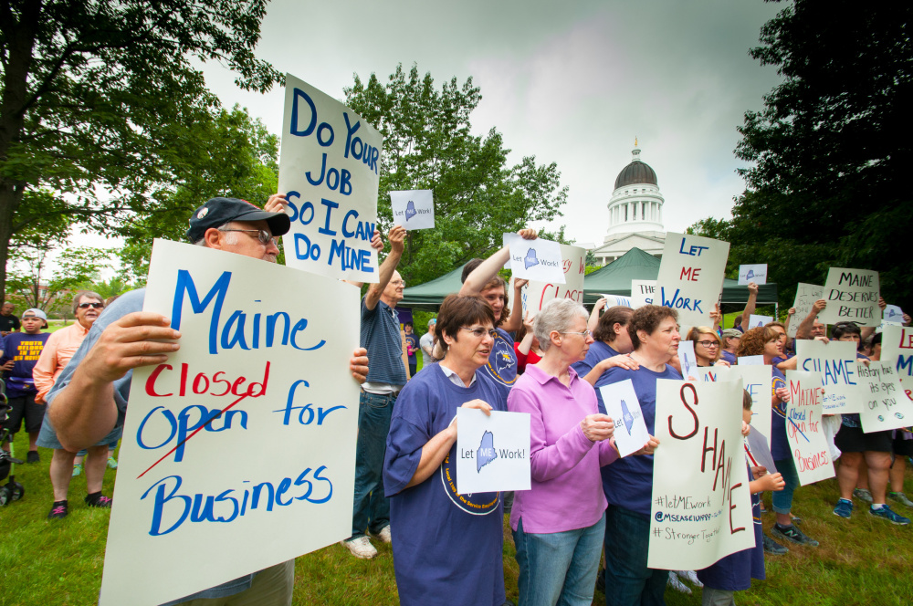 Protesters hold signs Saturday morning in Capitol Park in Augusta, in front the the State House, objecting to the state government shutdown that resulted from lawmakers' failure Friday to pass a budget that was veto-proof or acceptable to Gov. Paul LePage.