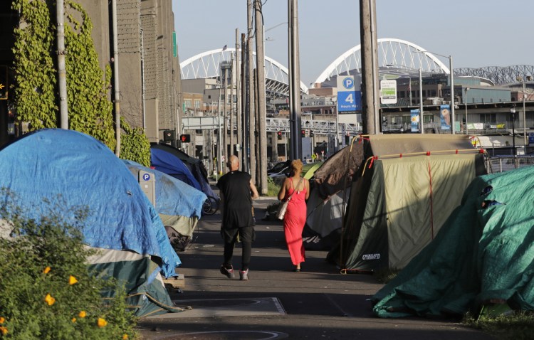 Two people walk past a half-dozen tents set up along a sidewalk at the Seattle waterfront. The city's latest tax proposal would hit Amazon the hardest, requiring employers making at least $20 million in gross revenues to pay $500 a year per worker.